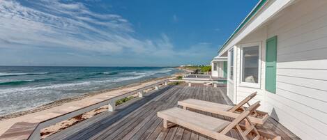 Une terrasse en bois avec des chaises longues, un bâtiment blanc aux volets verts et une vue sur la plage.