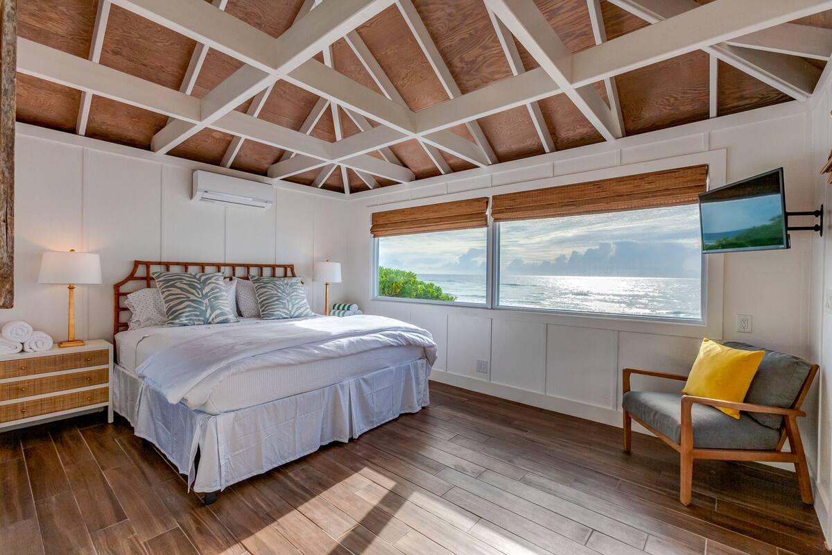 Un dormitorio con techo de madera, una cama con cubrecama blanco, una mesita de noche con lámpara, un sillón con cojín y una ventana con vista al mar.