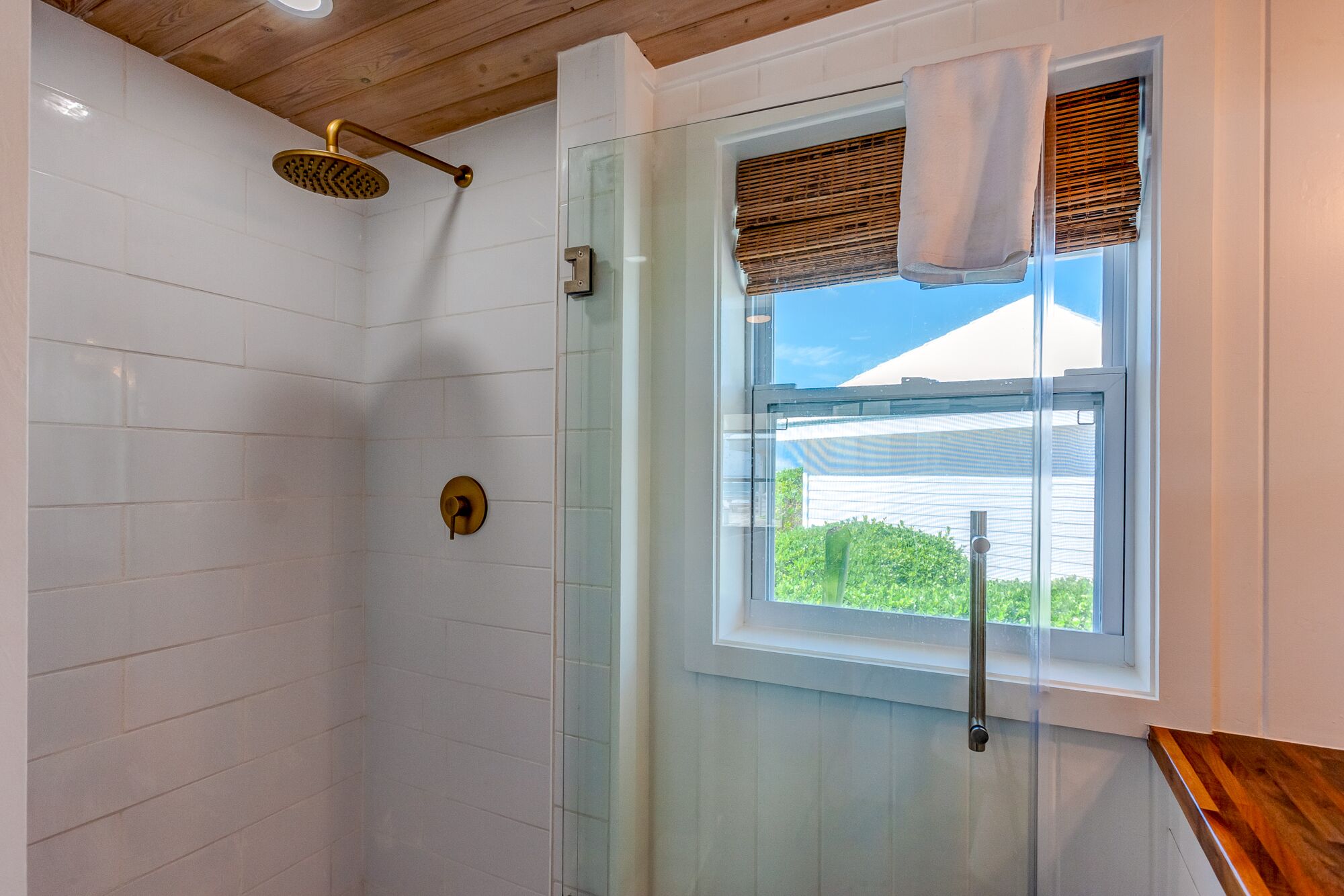 A shower with a glass door, a window with a towel hanging on the sill, and a wooden ceiling.