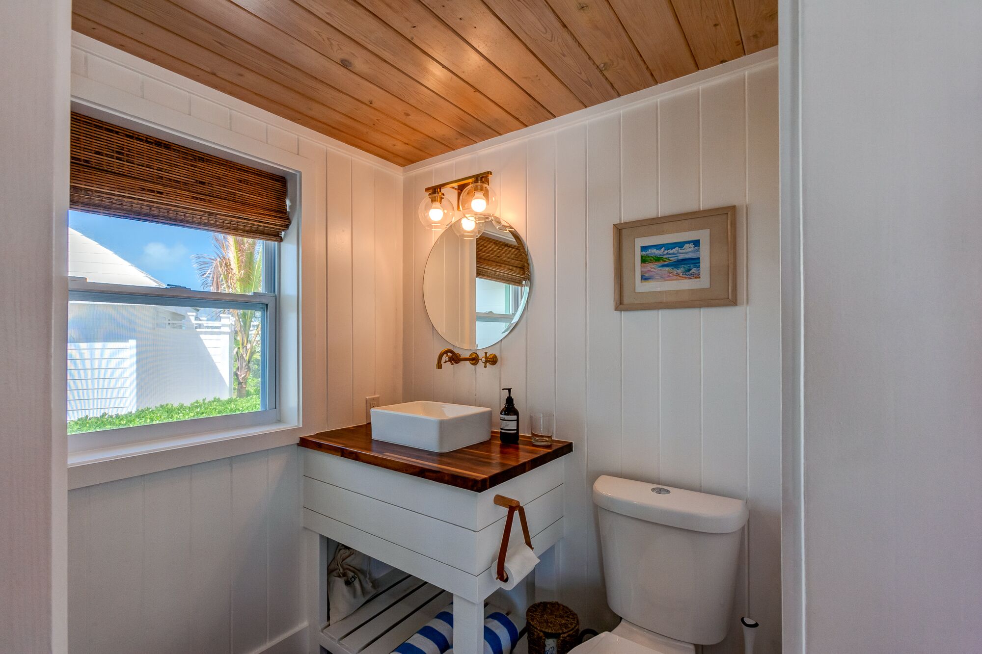 A bathroom with a wooden ceiling, a window with blinds, a white sink with a mirror above it, and a framed picture on the wall.