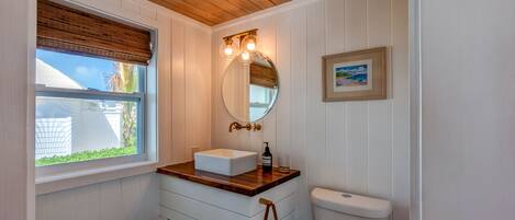 A bathroom with a wooden ceiling, a window with blinds, a white sink with a mirror above it, and a framed picture on the wall.