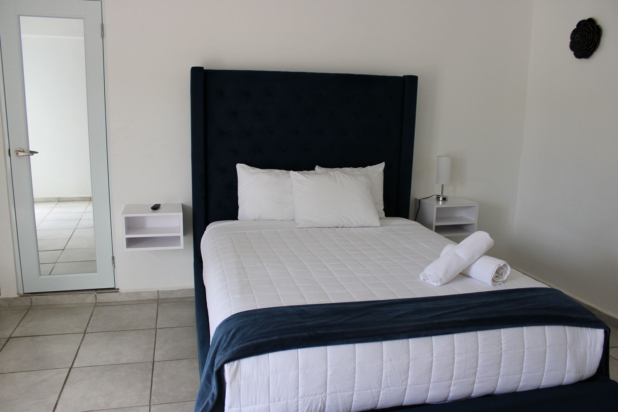 Una habitación de hotel con una cama grande, ropa de cama blanca, un cabecero oscuro, una mesita de noche con una lámpara y un pequeño estante.