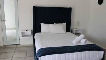 Una habitación de hotel con una cama grande, ropa de cama blanca, un cabecero oscuro, una mesita de noche con una lámpara y un pequeño estante.
