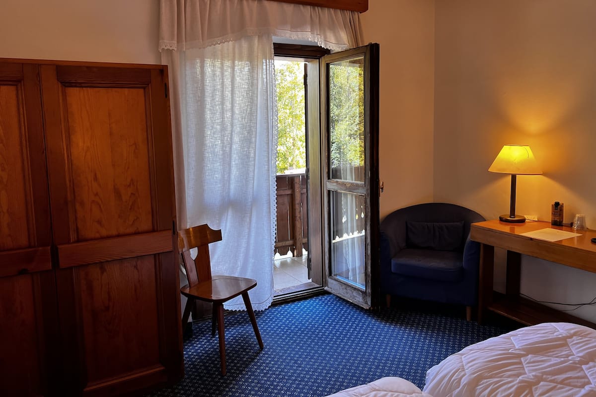 A hotel room with a wooden wardrobe, a chair, a desk with a lamp, and a bed.