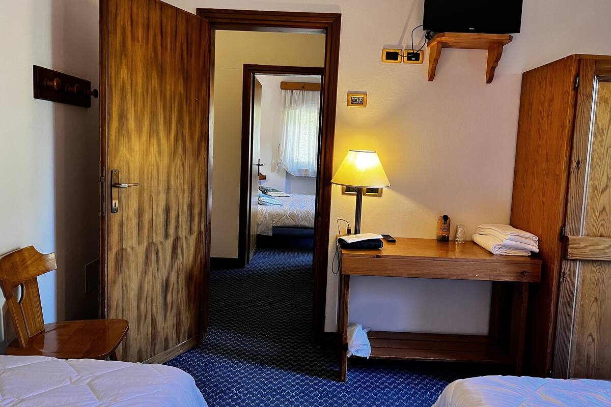 A hotel room with a wooden door, a bed with white bedding, a desk with a lamp, a TV mounted on the wall, and a small table with towels.