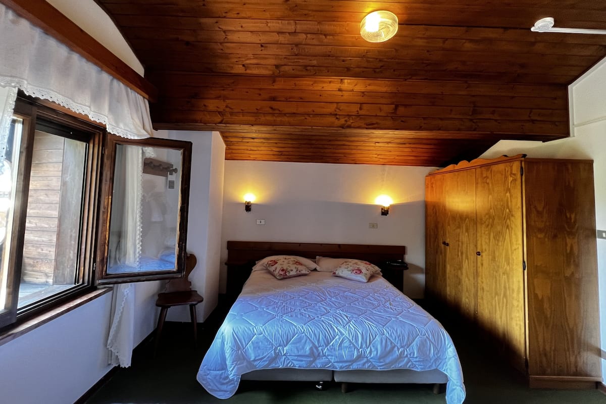 A bedroom with a wooden ceiling, a bed with a white quilt, two wall-mounted lights, a wooden wardrobe, and a window with curtains.