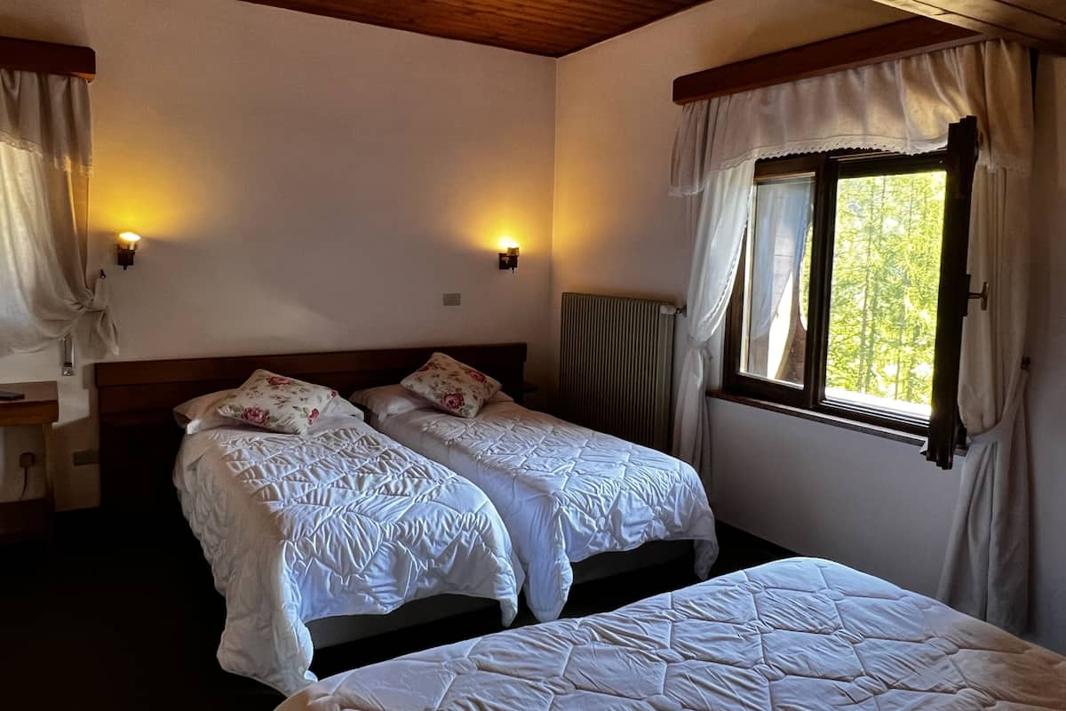 A room with two beds, wooden ceiling beams, a radiator, and a window with curtains.