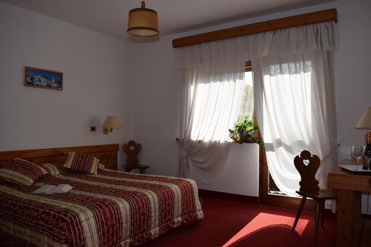 A bedroom with a bed, two bedside tables, a window with curtains, a small table with a lamp, and a chair.