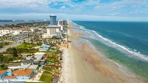 Vue aérienne d’une zone côtière avec une plage, des bâtiments et un ciel dégagé.