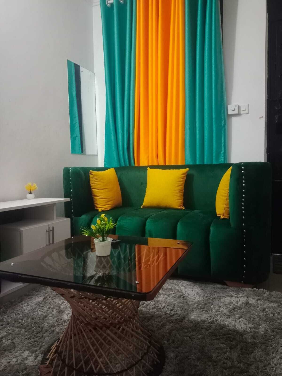 Ein Wohnzimmer mit einem grünen Sofa, gelben Kissen, einem Glastisch und einem gestreiften Vorhang.