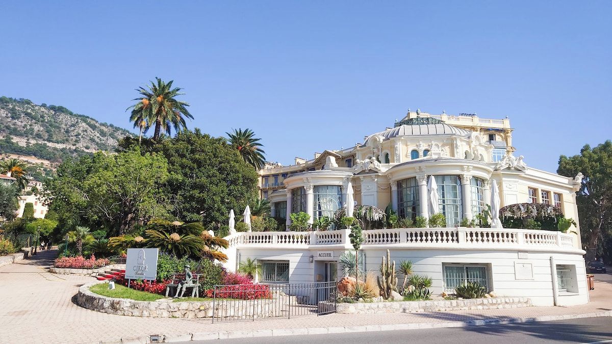Un edificio grande y ornamentado con fachada curva, varios balcones y un jardín ajardinado con palmeras y flores rojas.