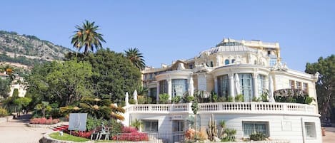 A large, ornate building with a curved facade, multiple balconies, and a landscaped garden with palm trees and red flowers.