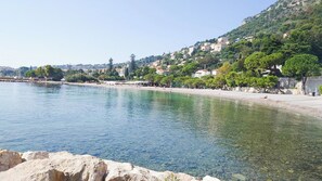 A coastal view with clear water, a sandy beach, and buildings on a hillside.