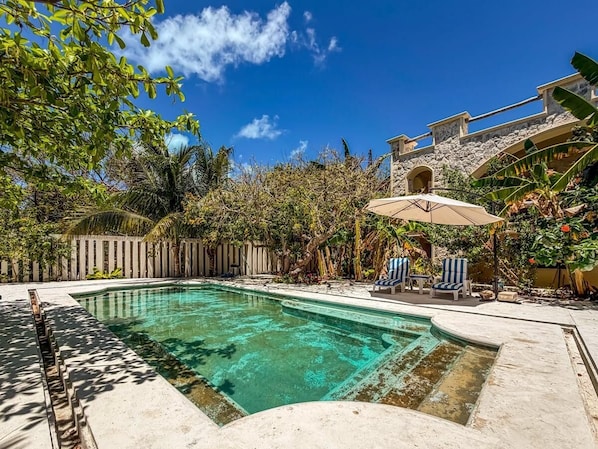 Une piscine dotée d’une plage en pierre, entourée de plantes tropicales et d’une clôture en bois.