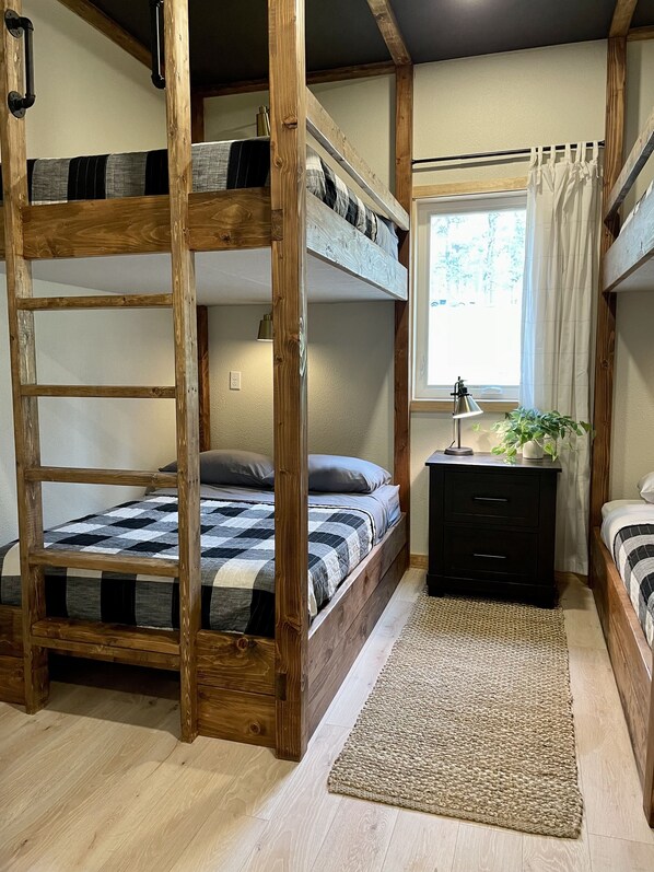 A bunk bed room with a wooden ladder, a small desk with drawers, a window with curtains, and a woven rug on the floor.