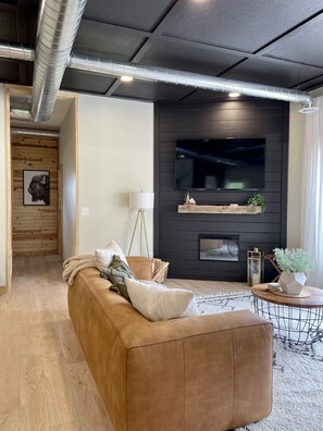 A modern living room with a brown leather sofa, a flat-screen TV mounted on a dark wall, and a wooden floor.