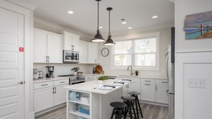 A modern kitchen with white cabinetry, a central island with stools, a microwave, and a clock on the wall. - 4 Luxury Home w/ Pool, FirePit, Outdoor Dining,| The Marshes Broad View (Hilton Head Island)