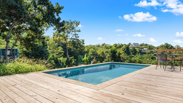 A rectangular swimming pool on a wooden deck with a view of a lush green landscape and a clear blue sky.