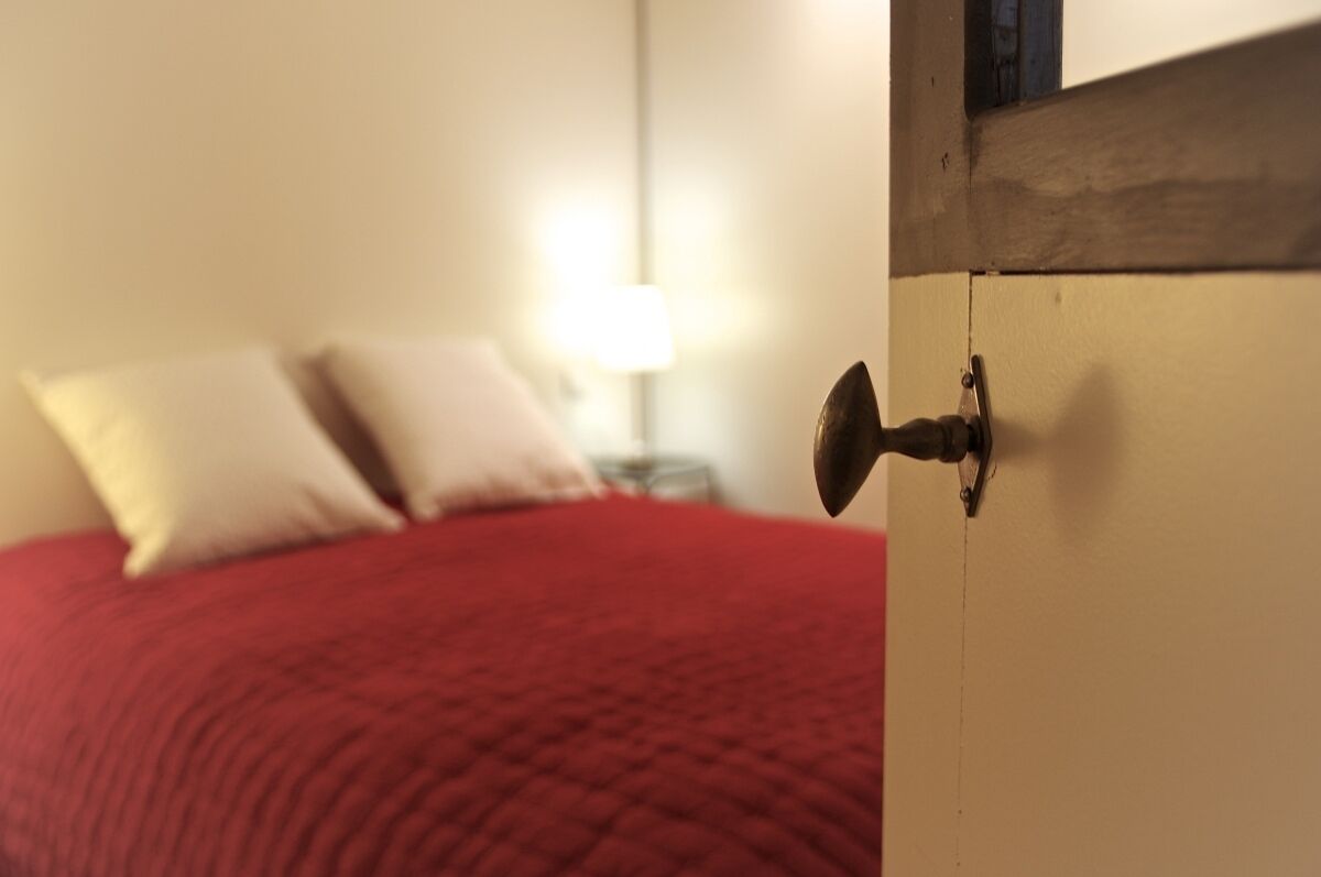 A bedroom with a red bedspread, a white wall, and a nightstand with a lamp.