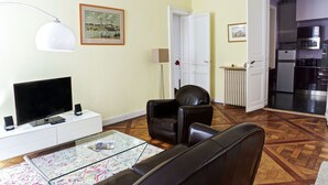 A living room with a flat-screen TV, a glass coffee table, a black leather sofa, a white cabinet, and a door leading to a kitchen. - Louis Combes - Localisation Exceptionnelle / 1 cha (Burdeos)