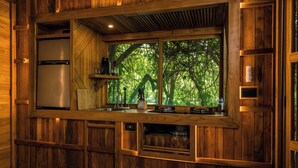 Een houten keukentje met een inbouwkoelkast, een koffiezetapparaat en een raam met uitzicht op een bos.