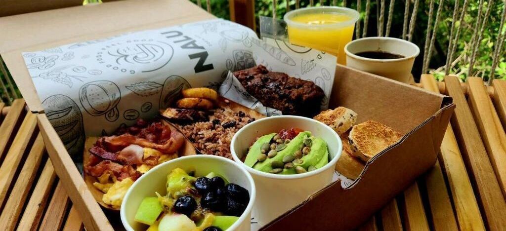 A meal tray with various breakfast items including toast, bacon, eggs, and fruit, served on a wooden table outdoors.