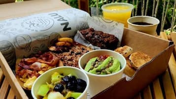 A meal tray with various breakfast items including toast, bacon, eggs, and fruit, served on a wooden table outdoors.