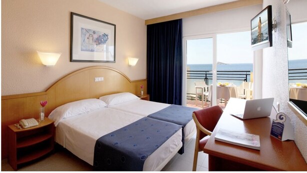 A hotel room with a bed, a desk with a laptop, a chair, a nightstand with a phone, and a view of the sea through large windows. - Hotel Tiburon (Guadalajara)