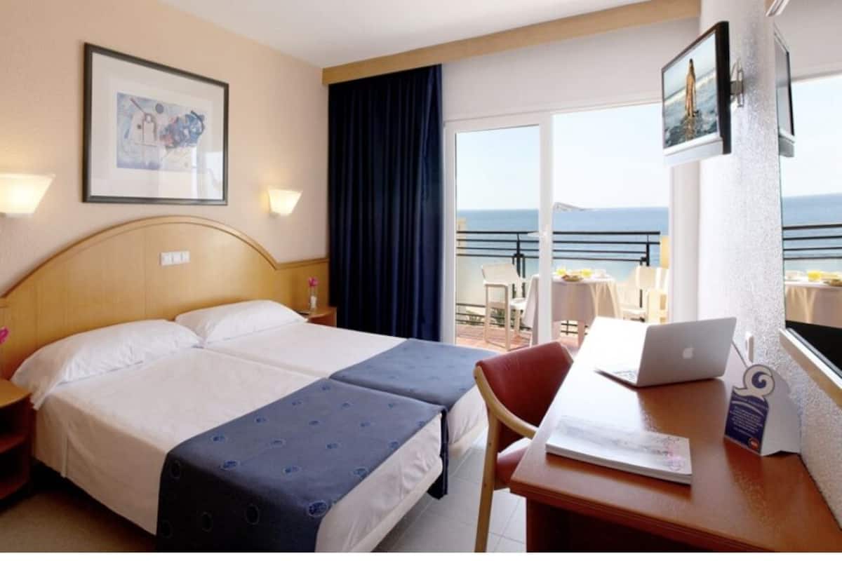 A hotel room with a bed, a desk with a laptop, a chair, a nightstand with a phone, and a view of the sea through large windows.