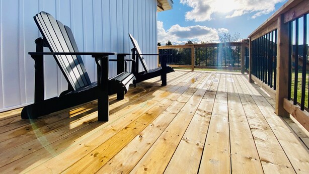 A wooden deck with Adirondack chairs against a white building, overlooking a grassy area. - Cottage #6 - 1 bedroom - pet friendly! (French River)