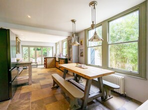 Une cuisine avec une table et un banc en bois, de grandes fenêtres donnant sur un paysage verdoyant, et un radiateur placé sous la fenêtre.