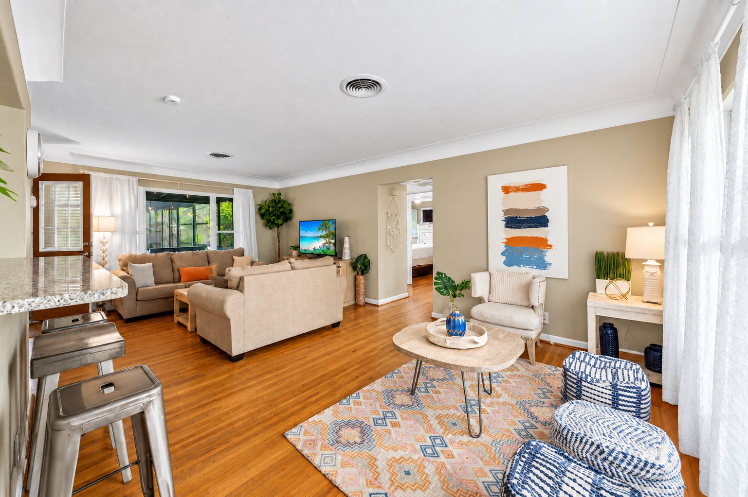 A modern living room with a sofa, coffee table, and a TV. There is a dining area with a bar and stools. The room has wooden flooring and a large window.