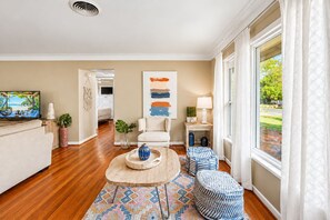 Un salon moderne comprenant un téléviseur, un canapé, une table basse et des poufs. Un tableau est accroché au mur et il y a une fenêtre avec des rideaux.