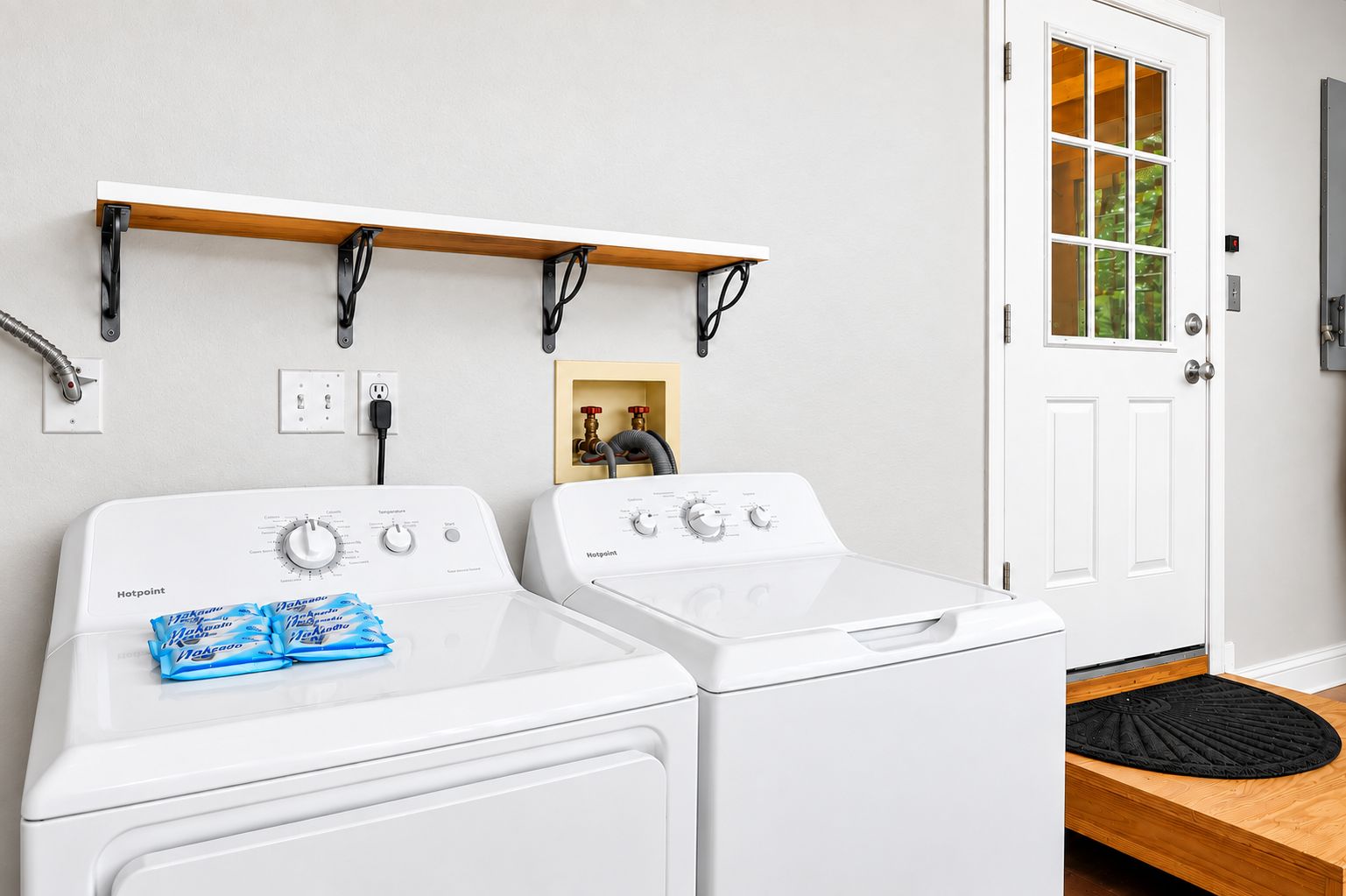 A laundry room with two white washing machines, a wooden shelf above, and a door with a window.