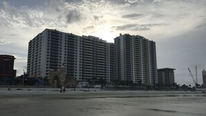 A high-rise building with multiple balconies, a beach in the foreground, and a cloudy sky.