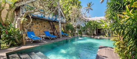 A clear pool with blue lounge chairs, surrounded by lush greenery and a stone building with a blue railing.