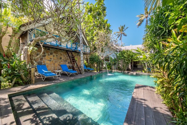 A clear pool with blue lounge chairs, surrounded by lush greenery and a stone building with a blue railing.
