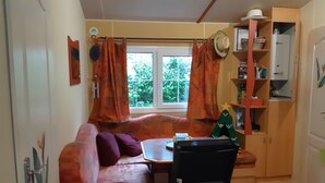 A cozy room with a sofa, a small table, and a chair. There is a window with curtains, a shelf with hats, and a door.