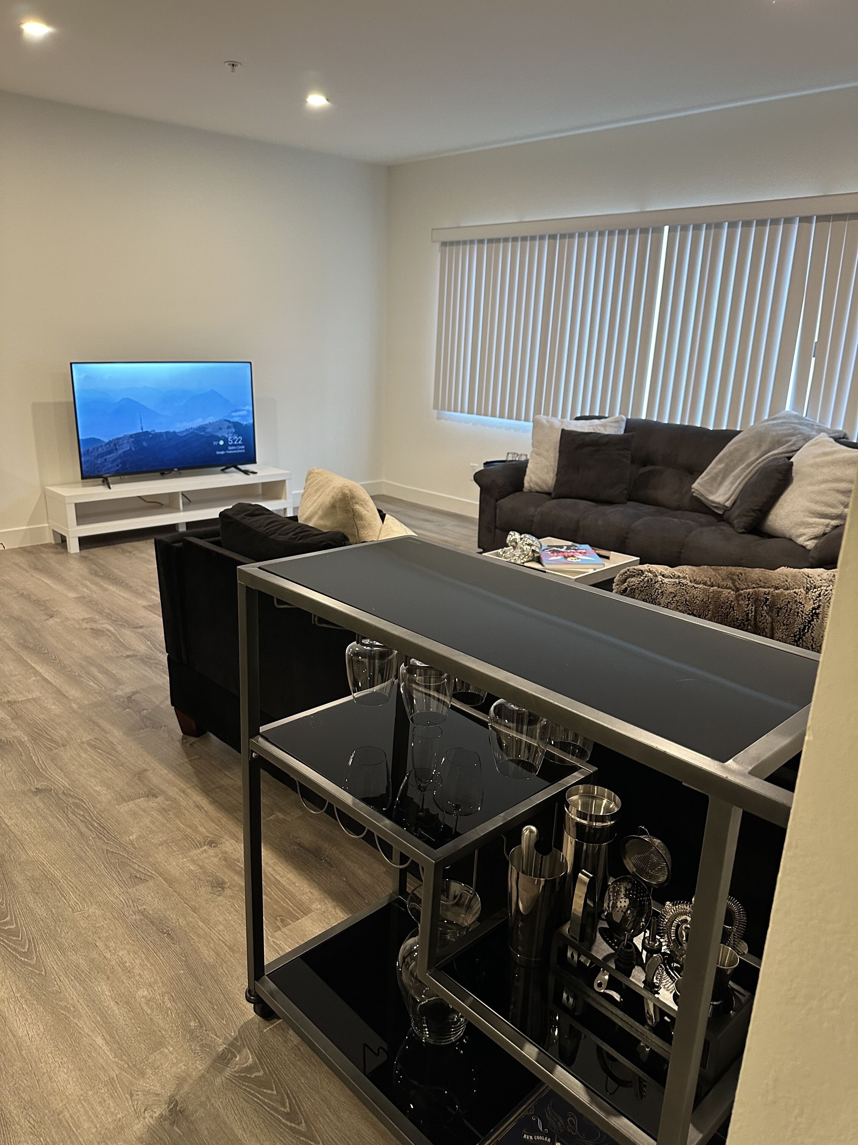 A modern living room with a flat-screen TV, a sofa, a coffee table, and a glass-top dining table with wine glasses and a coffee machine.