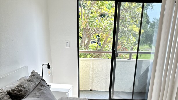 A bedroom with a bed, a nightstand, a lamp, and a view of a balcony with trees.