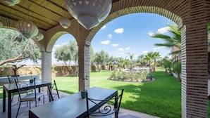 An outdoor patio with tables and chairs, a brick archway, and a view of a well-maintained lawn and palm trees. - Villa Chems (Marrakech)