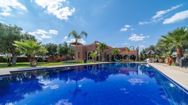 A swimming pool with a clear blue water surface, surrounded by palm trees and a building with arched walkways. - Villa Chems (Marrakech)