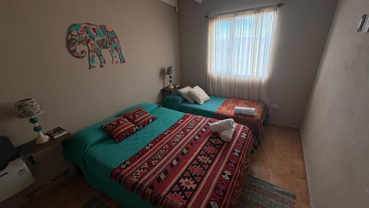 A room with two beds, a wall decoration, a window with curtains, and a small table with a lamp.