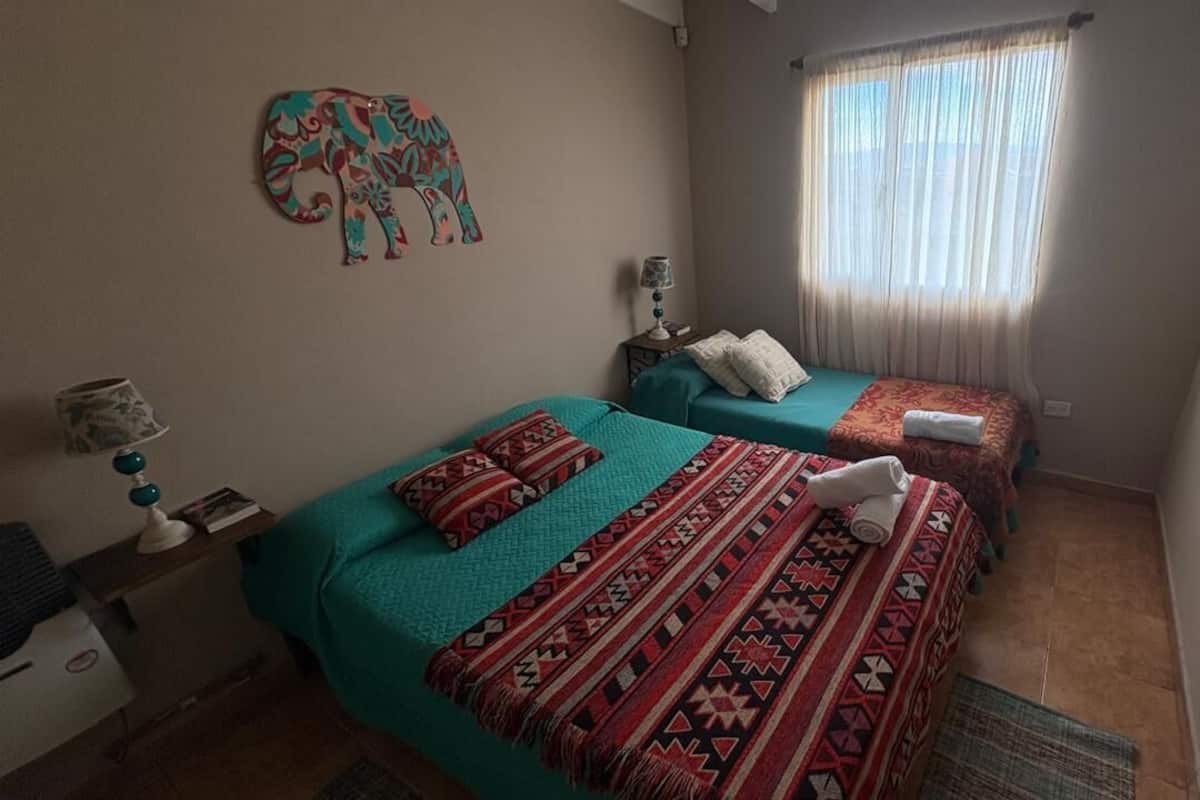 Una habitación con dos camas, un adorno en la pared, una ventana con cortinas y una mesita de noche con una lámpara.