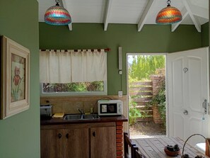 Cocina con microondas, pileta y alacenas de madera. Hay una ventana con cortina y una puerta que da al exterior.