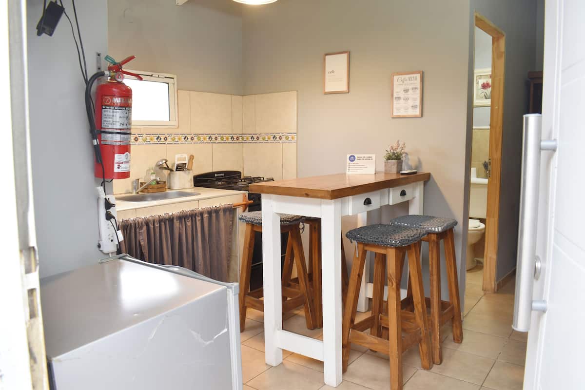 Una cocina compacta con refrigerador, estufa, taburetes de madera, extintor de incendios y un cartel con la carta de café.