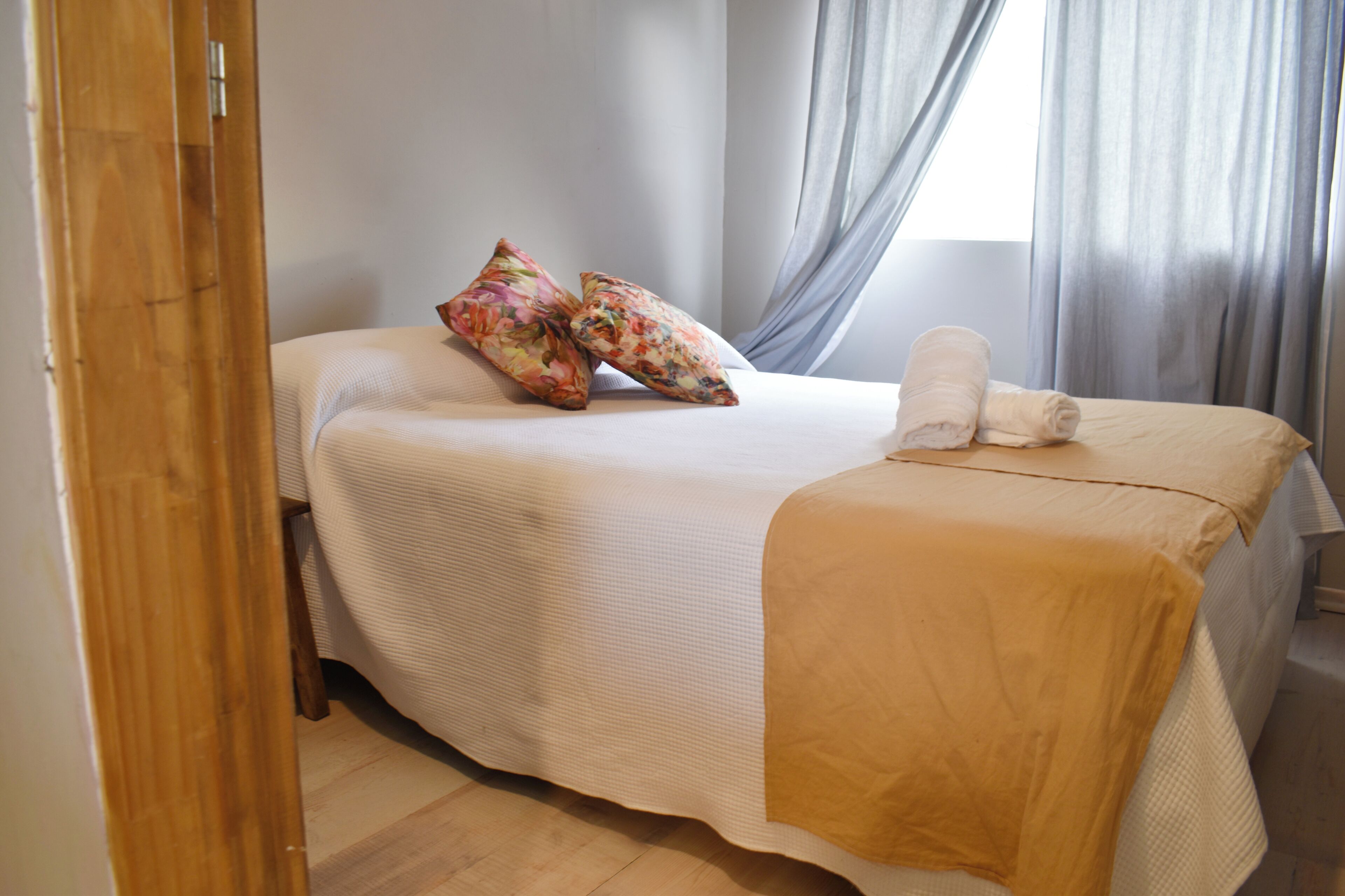 Una cama bien hecha con una colcha blanca y una manta color beige, dos almohadas estampadas con motivos florales y una toalla enrollada al lado.