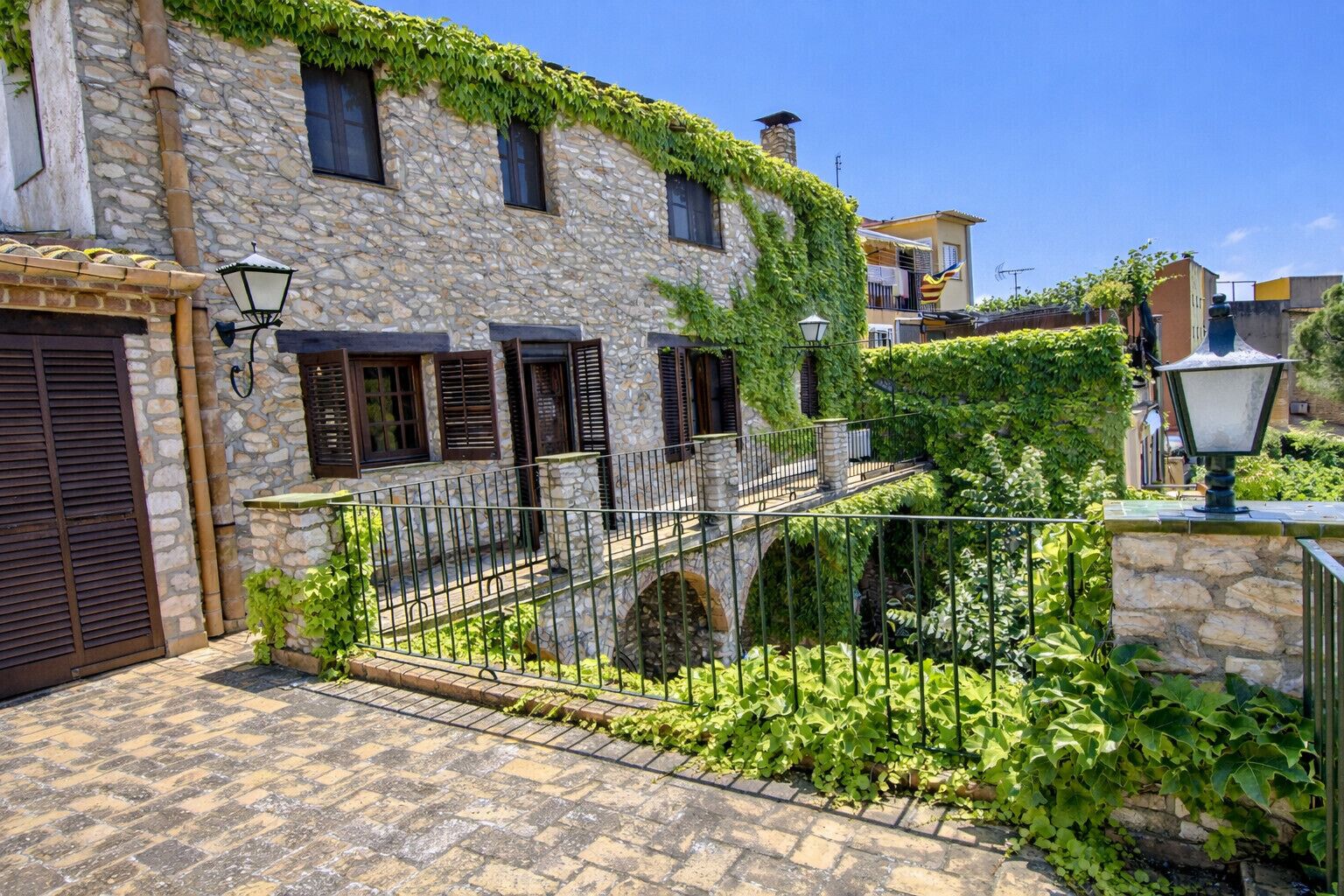 Un edificio de piedra con contraventanas, una cerca de metal y un patio empedrado.