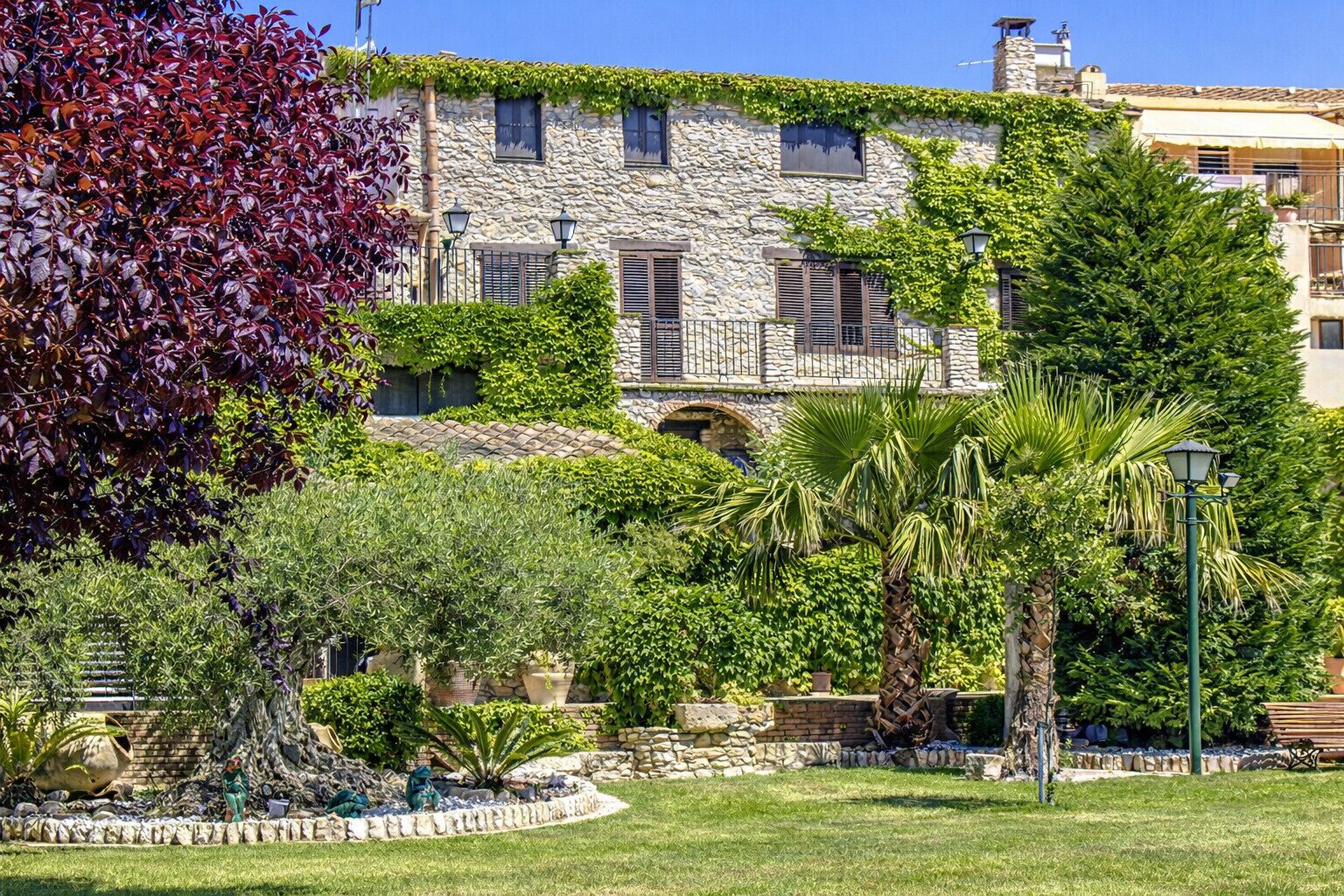 Un edificio de piedra con un jardín delantero, que incluye una palmera y varios arbustos.
