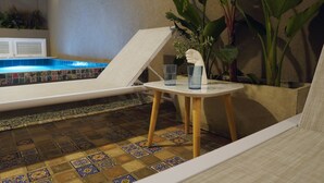 Area piscina con pavimento piastrellato, sedie a sdraio, un tavolino con un vaso e bicchieri, e una parete decorata con piante verdi.